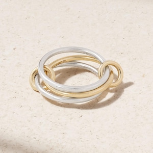 May include: A three-ring interlocking ring in silver and gold. The ring features two silver bands and one gold band, with the gold band marked "18K". The rings are interlocked, creating a unique design.