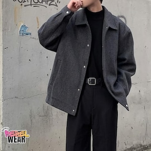 May include: A charcoal gray collared jacket with a button-front closure, long sleeves, and side pockets, worn over a black mock-neck shirt and black trousers with a black belt featuring a silver square buckle. The jacket has a textured fabric finish.
