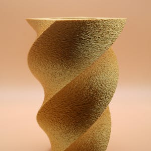 Könnte beinhalten: Eine strukturierte, goldgelbe Vase mit einem gedrehten, spiralförmigen Design. Die Vase hat eine breite Öffnung oben und eine strukturierte Oberfläche. Der Hintergrund ist einfarbig pfirsichfarben. Die Vase ist etwa 25 cm hoch.