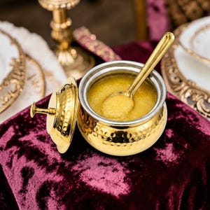 May include: A gold-colored serving bowl with a lid and spoon, filled with a yellow substance. The bowl has a hammered texture and a silver interior. The spoon rests inside the bowl. The lid is placed next to the bowl on a burgundy velvet surface.