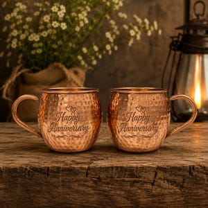 Pode incluir: Duas canecas de cobre estilo Moscow Mule com textura martelada e alças curvas. Cada caneca tem as palavras "Happy Anniversary" gravadas na frente, com detalhes de corações e arabescos. As canecas estão sobre uma superfície de madeira.