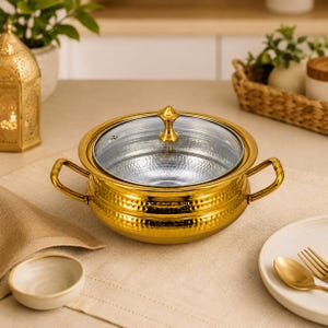 Könnte beinhalten: Eine runde, goldfarbene Servierschale mit gehämmertem Äußeren, zwei seitlichen Griffen und einem klaren Glasdeckel mit goldfarbenem Knauf. Das Innere der Schale ist silberfarben und metallisch, was einen Kontrast zum Goldfinish bildet.