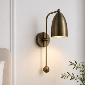 Può includere: Una lampada da parete con finitura in ottone spazzolato, dotata di una base circolare, un lungo braccio verticale e un paralume a forma di cupola che dirige la luce verso il basso. L'apparecchio include un piccolo elemento decorativo.