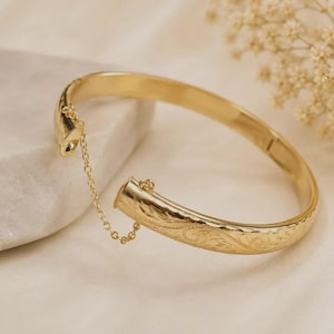 May include: A gold-toned hinged bangle bracelet featuring an intricate engraved scrollwork pattern across its surface. The bracelet includes a delicate attached safety chain that connects the two ends, ensuring the piece remains secure when opened.