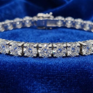 May include: A silver-toned tennis bracelet featuring a continuous row of round, clear, faceted stones set in individual metal prongs. The bracelet includes a secure box clasp mechanism at the center, showcasing a uniform design of sparkling circular gems.