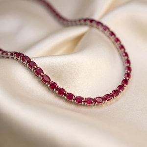 May include: A tennis necklace featuring a continuous row of oval-cut, deep red rubies set in a polished silver-toned metal frame. Each individual gemstone is secured by four small prongs, creating a flexible and uniform chain design.