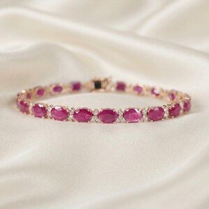 May include: A tennis bracelet featuring a series of oval-cut, deep pinkish-red rubies alternating with small, clear, round-cut accent stones set in a polished metal band with a secure clasp mechanism visible at the center of the circular arrangement.