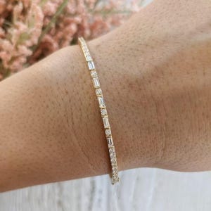 Pode incluir: Uma delicada pulseira de ouro com um padrão repetitivo de pedras claras alternadas em corte baguette e redondo, engastadas numa banda de metal fina. O design cria uma linha contínua de brilho que envolve o pulso com elegância.