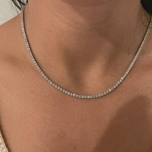 May include: A silver-toned tennis necklace featuring a continuous row of small, round, clear faceted stones set in a metallic chain. The necklace rests against the neck, showcasing a uniform design of sparkling stones linked together in a single strand.