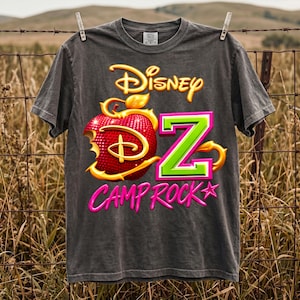 May include: A charcoal gray short-sleeve t-shirt featuring a graphic print with the text Disney, a large red bitten apple with gold accents, a bright green letter Z, and the pink text CAMP ROCK with a small pink star at the end of the word.
