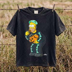 May include: A black short-sleeve t-shirt featuring a graphic of a yellow cartoon character wearing a skeleton costume with light blue bones, a teal skull mask, and holding an orange jack-o'-lantern candy bucket while eating a chocolate treat.