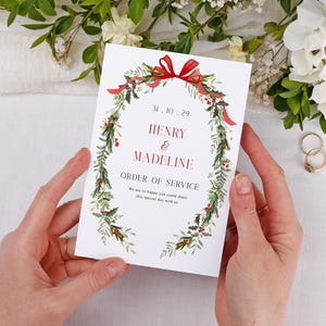 May include: A white wedding order of service card held by hands, with a green and red wreath design. The card displays the date, names "Henry & Madeline", and "Order of Service". Two gold wedding rings are visible in the background.