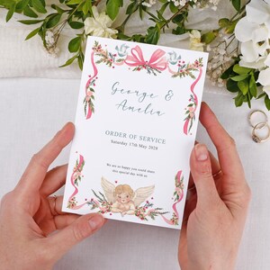 May include: A wedding order of service card held by hands. The card features the names "George & Amelia" and the date "Saturday 17th May 2028". It is decorated with pink ribbons, floral accents, and a cherub illustration.