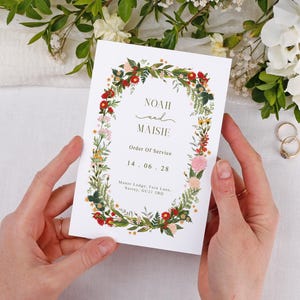Peut inclure: Un faire-part de mariage blanc, tenu par des mains, avec les noms "Noah et Maisie" et la date "14.06.28". Il est orné d'une couronne florale avec des fleurs rouges, jaunes et roses. Deux alliances dorées sont visibles en arrière-plan.