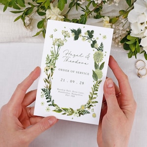 May include: A wedding order of service card with a green floral design and the names "Hazel & Theodore". The date "21.09.28" is displayed. The card is held by hands, with wedding rings and flowers in the background.
