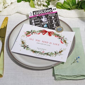 May include: A wedding invitation with the text "ALL YOU NEED IS LOVE AND A BIT OF LUCK" sits on a plate with a lottery ticket and a coin. A gold fork and knife are on the left, and a green napkin is on the right.