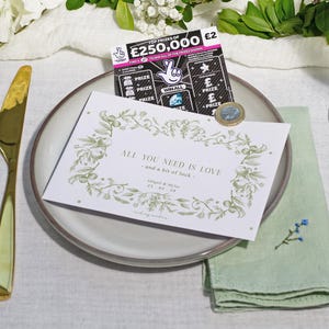 Peut inclure: Mise en place de mariage avec une carte blanche portant l'inscription "ALL YOU NEED IS LOVE" et des motifs floraux. Un billet de loterie, une pièce de monnaie, des couverts dorés et une serviette verte sont également présents.