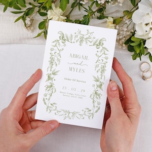 May include: A wedding order of service card held in two hands, with the names "ABIGAIL and MYLES" in a decorative font. The card has a green floral design and the date "23.03.29". The card is surrounded by flowers and wedding rings.