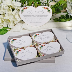 May include: A gray box holding heart-shaped cards with floral designs and text such as "Sp & Say," "Most Likely," "Two Truths and a Lie," "Compliment Roulette," and "Photo Challenge." A champagne glass is visible in the background.