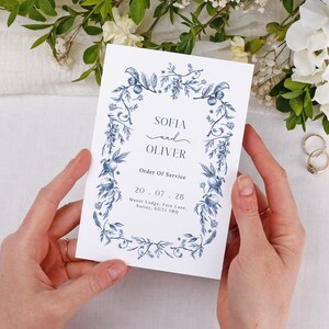 May include: A white wedding program with a blue floral design, held by hands. The program displays the names "SOFIA and OLIVER", the date "20.07.28", and "Order Of Service". Wedding rings and flowers are visible in the background.