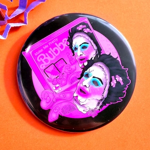 May include: A round black pin-back button featuring a vibrant pink graphic of two faces with stitched skin masks, dark hair, and blue eye makeup. The design includes a box labelled Make Me Pretty Bubba with various beauty accessories.