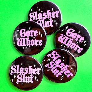 May include: A collection of five circular pin-back buttons with a glossy black finish. The buttons feature light pink text reading either Slasher Slut or Gore Whore, accented by small white four-pointed star graphics scattered around the lettering.