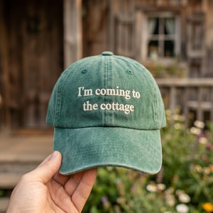 May include: A green baseball cap with the phrase "I'm coming to the cottage" in white lettering. The hat is made of a soft material and has a curved brim. The background is a blurred view of a wooden building and foliage.