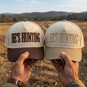 May include: Two baseball caps with the text HE'S HUNTING embroidered on the front. The left cap is cream with a dark brown brim and dark brown lettering. The right cap is cream with a matching cream brim and tonal cream lettering.