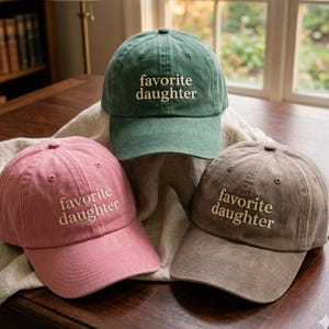 May include: Three baseball caps in green, pink, and brown, each featuring the embroidered text "favorite daughter" in a light-colored thread. The caps have a structured crown, a curved brim, and a washed fabric finish with visible stitching.