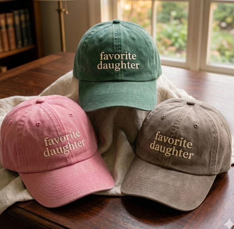 May include: Three baseball caps in green, pink, and brown, each featuring the embroidered text "favorite daughter" in a light-colored thread. The caps have a structured crown, a curved brim, and a washed fabric finish with visible stitching.