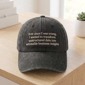 May include: A charcoal gray baseball cap featuring embroidered beige text that reads: Ever since I was young, I wanted to transform unstructured data into actionable business insights. The cap has a six-panel construction with a curved brim.