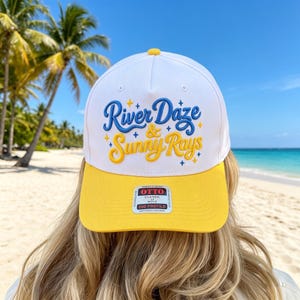 May include: A white baseball cap with a yellow brim featuring the embroidered blue and yellow text River Daze & Sunny Rays with small star accents. A rectangular sticker on the brim reads Otto Classic 550 Profile in black and white text.