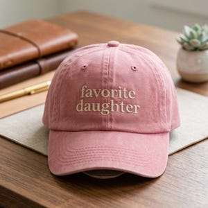 May include: A dusty rose baseball cap featuring the embroidered text favorite daughter in a light beige serif font. The cap has a structured crown, a curved brim, and a button on top, constructed from a soft, matte fabric with visible stitching.