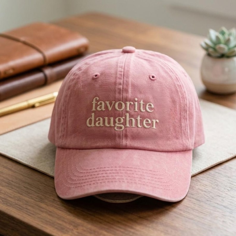 May include: A dusty rose baseball cap featuring the embroidered text favorite daughter in a light beige serif font. The cap has a structured crown, a curved brim, and a button on top, constructed from a soft, matte fabric with visible stitching.
