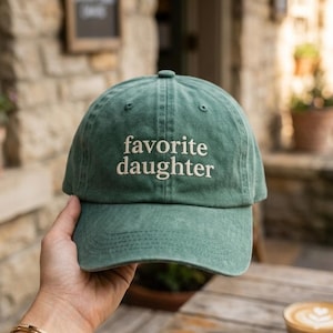 May include: A washed green baseball cap featuring the embroidered text "favorite daughter" in a cream-colored serif font across the front panels. The cap has a curved brim, a button on the top, and visible stitching details along the crown and brim.