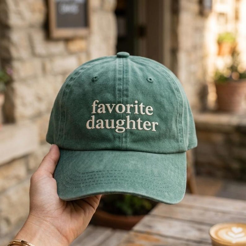 May include: A washed green baseball cap featuring the embroidered text "favorite daughter" in a cream-colored serif font across the front panels. The cap has a curved brim, a button on the top, and visible stitching details along the crown and brim.