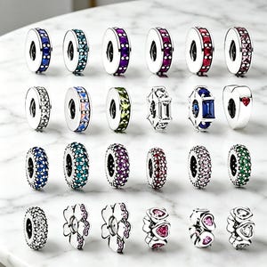 May include: A collection of silver charm beads with various colorful gemstone accents. The beads feature designs with blue, green, purple, red, and pink stones, as well as heart and flower shapes. The charms are displayed on a white marble surface.