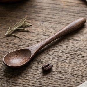 May include: A wooden spoon with a rounded handle and oval bowl rests on a wooden surface. A coffee bean and a sprig of rosemary are also on the surface. The spoon is a dark brown color.