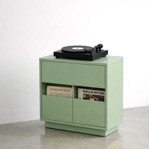 Può includere: Un mobile verde menta per dischi in vinile con un giradischi nero sopra. Il mobile ha due scomparti aperti con dischi in vinile. Gli album sono etichettati "ECHOS OF GOLD" e "MODULAR RHYTHMS".