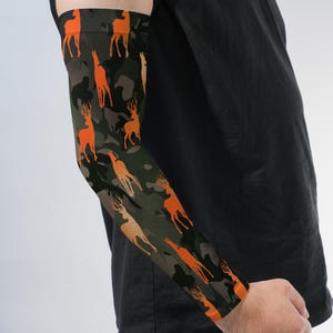 May include: An arm sleeve featuring a camouflage pattern in shades of dark green, black, and brown, overlaid with a repeating print of bright orange deer silhouettes. The sleeve is fitted to the arm and extends from the upper bicep to the wrist.