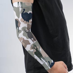 May include: An arm sleeve featuring a camouflage pattern in shades of white, tan, and olive green. The fabric is decorated with dark navy blue silhouettes of wild turkeys and deer with antlers, arranged in a repeating graphic print design.