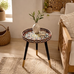 May include: A round side table with three black legs featuring gold-toned tips. The tabletop is a circular tray with a colorful, intricate mandala-style pattern in shades of red, green, and yellow. A small ceramic vase and a glass sit on top.