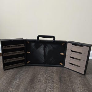 May include: A black portable storage case with a top handle, featuring a central compartment and two side sections. The left side has four dark brown open shelves, while the right side contains four light beige drawers with angled front panels.