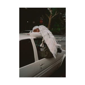 May include: A person wearing a white head covering and a white shirt, leaning on a white car. A large white feathered wing is attached to the car. The background includes a street, trees, and a stop sign.