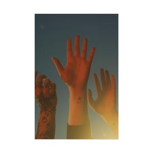 May include: Three arms are raised against a blue sky, bathed in warm sunlight. One arm has a tattoo that says "HAR" and a floral design. Another arm has a small tattoo of a bottle. The image evokes a sense of freedom and connection.