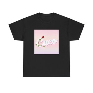 May include: Black t-shirt featuring a square graphic with the word "Cuco" in a cursive font, adorned with a red rose and green vines. The graphic has a pink and white gradient background. The shirt is made of a soft material.