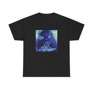 May include: Black t-shirt featuring a blue-toned image of a person wearing sunglasses and holding small objects. The image has a surreal, dreamlike quality. The t-shirt is a classic crew neck style.