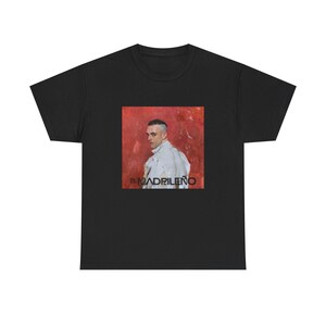 Puede incluir: Camiseta negra con una imagen cuadrada de un hombre con una chaqueta blanca sobre un fondo rojo. La imagen tiene el texto "EL MADRILEÑO" en negro. La camiseta es de cuello redondo.