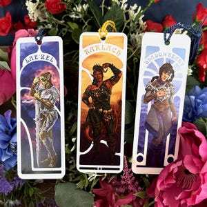 May include: Three fantasy character bookmarks with colorful illustrations. Each bookmark features a different character with a name at the top: Lae'zel, Karlach, and Shadowheart. The bookmarks have a white border and a tassel at the top.