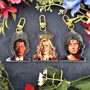 May include: Three clear acrylic keychains featuring portraits of individuals with gold-colored hardware. The portraits have a painted appearance, with one wearing a crown. The keychains are surrounded by floral decorations.
